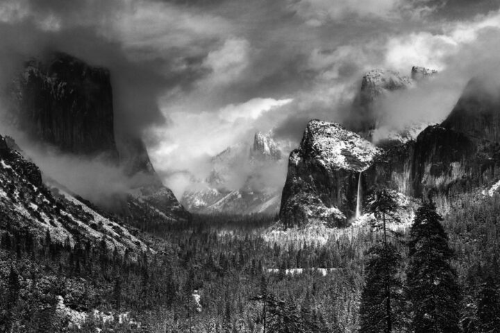 Black & white landscape photography by Ansel Adams. Clouds above a snow, capped valley and forests in Yosemite National Park, USA